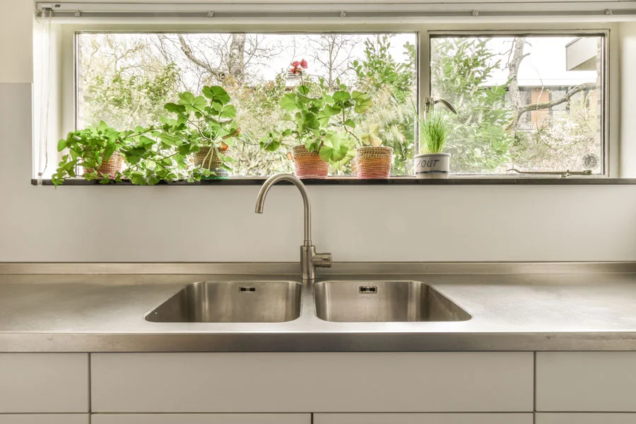 A kitchen sink and a window with plants in the wicker of the window facing the street
